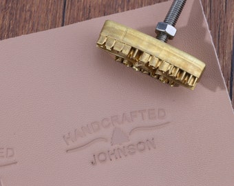 Könnte beinhalten: Ein Stempelwerkzeug aus Messing zum Prägen von Leder mit einem Griff und einem Design, das "HANDCRAFTED JOHNSON" mit einer kleinen Baumgrafik zeigt. Das Werkzeug wird verwendet, um das Design in ein Stück hellbraunes Leder zu prägen.
