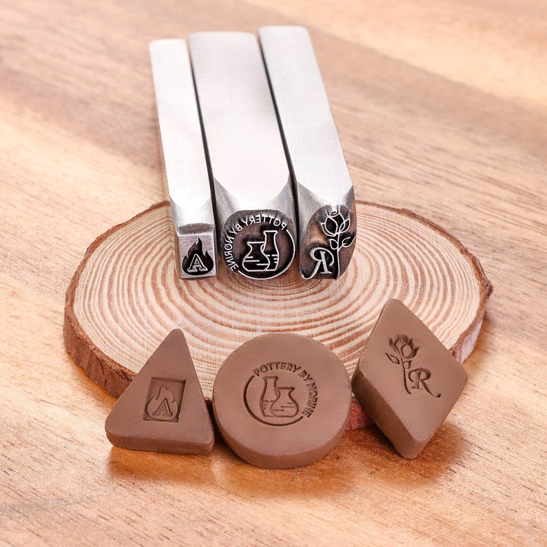 May include: Three metal stamps with different designs: a flame, a beaker, and a flower. The stamps are on a wooden surface with three clay impressions of the stamps.