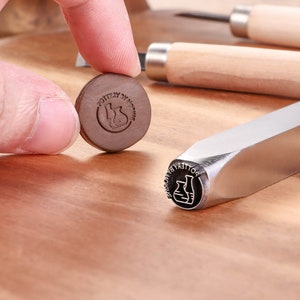May include: A close-up of a metal stamp with the text "POTTERY BY MOMO" and a graphic of two beakers. The stamp is on a wooden surface.