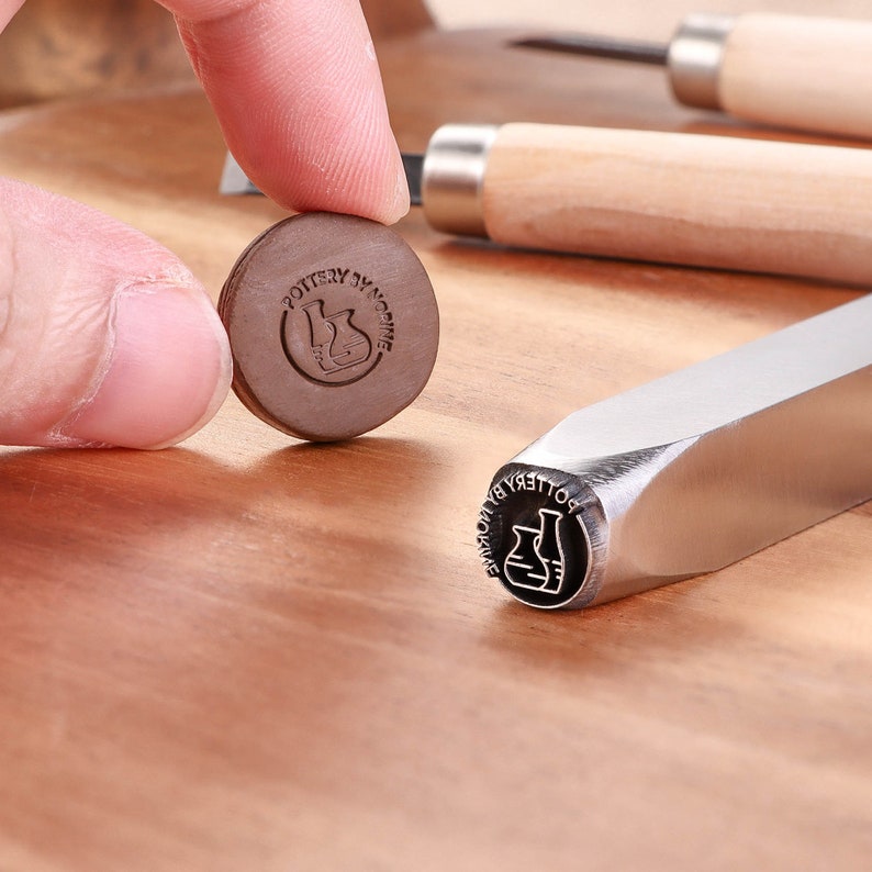 May include: A close-up of a metal stamp with the text "POTTERY BY MOMO" and a graphic of two beakers. The stamp is on a wooden surface.