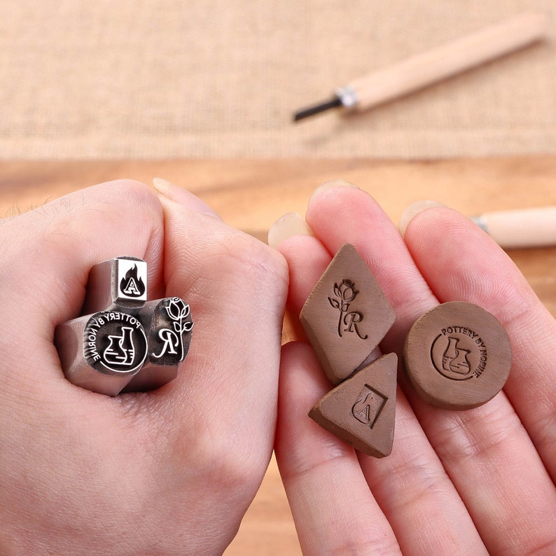 May include: A set of metal stamps for pottery, featuring a rose, a letter R, a beaker, and a flame. The stamps are held in a hand, along with three clay stamps with the same designs.
