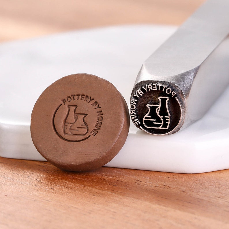 May include: A close-up of a metal stamp with the text "POTTERY BY NORINE" and a design of two pottery vases. The stamp is used to create impressions on clay.