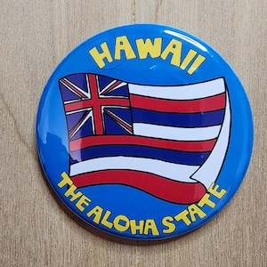 May include: A round, blue button featuring the Hawaiian flag with red, white, and blue stripes and a Union Jack in the upper left corner. The button has the words "HAWAII" and "THE ALOHA STATE" in yellow.