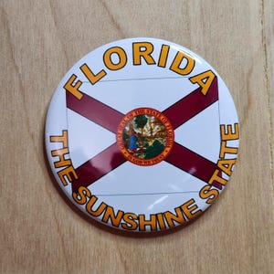 May include: A round button featuring the Florida state flag design. The button has a white background with a red cross and the state seal in the center. The words "FLORIDA" and "THE SUNSHINE STATE" are printed in orange around the edge.