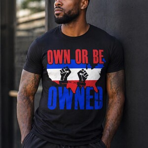 May include: Black t-shirt with the text "OWN OR BE OWNED" in red and blue. The design includes a white and blue map of the United States with two raised fists in black. The shirt is short-sleeved.