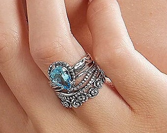 Puede incluir: Juego de anillos de plata con una piedra preciosa azul claro en forma de pera. El anillo presenta intrincados detalles de filigrana y está apilado con dos bandas adicionales. El diseño es ornamentado y elegante, adecuado para ocasiones especiales.