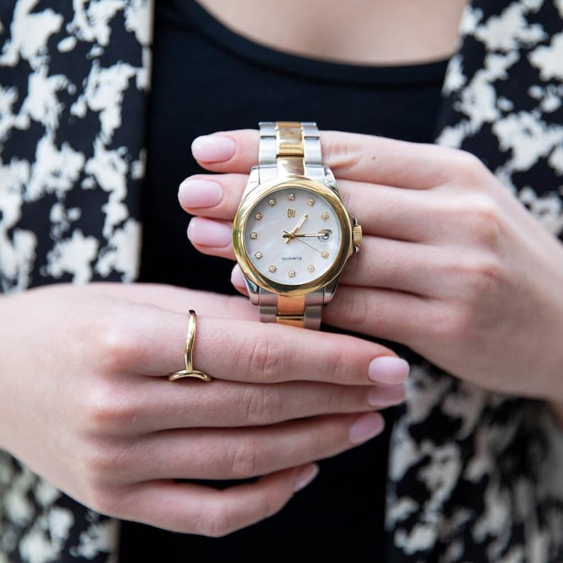 May include: A two-tone gold and silver watch with a white face and diamond accents. The watch has a silver band and a gold bezel. The watch face has a white background with black Roman numeral hour markers and a date window at the 3 o'clock position. The watch is set on a white background.