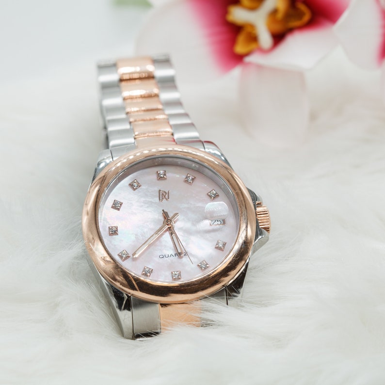 May include: A silver and rose gold tone watch with a white face and diamond accents. The watch face has a mother-of-pearl background and the word "QUARTZ" is printed on the face.