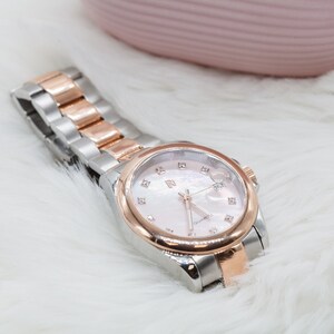 May include: A silver and rose gold watch with a white face and diamond accents. The watch has a quartz movement.