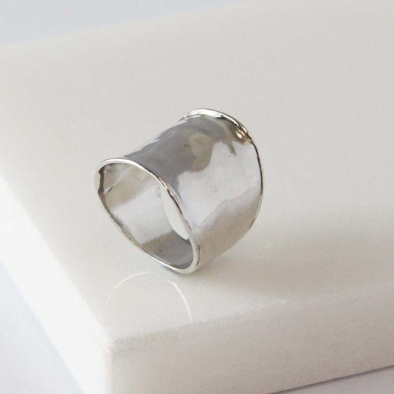 Puede incluir: Un anillo de plata con una banda ancha y martillada. El anillo es liso y tiene una forma ligeramente curvada.