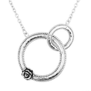 Puede incluir: Collar plateado con dos círculos entrelazados, uno adornado con una rosa. Los círculos tienen un borde texturizado y con cuentas, unidos por una fina cadena. El diseño es elegante y minimalista, perfecto para cualquier ocasión.