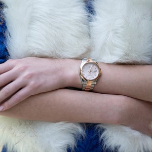 May include: A silver and rose gold wristwatch with a white face and black Roman numeral hour markers. The watch is on a woman's wrist.