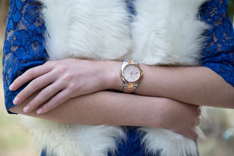 May include: A silver and rose gold wristwatch with a white face and black Roman numeral hour markers. The watch is on a woman's wrist.