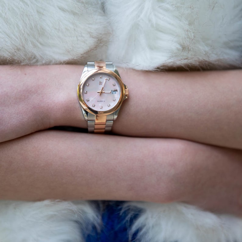 May include: A two-tone silver and rose gold watch with a white face and diamond accents. The watch has a silver band and a rose gold bezel.