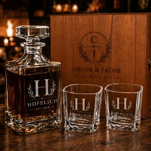 May include: A personalized whiskey decanter set with a wooden box. The set includes a square decanter, two square glasses, and a wooden box. The decanter and glasses are engraved with the letter "H" and the name "HOFELICH". The wooden box is engraved with the names "CHLOE & JACOB".