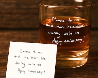 May include: A clear glass filled with a brown liquid, likely a spirit, with the words "Cheers to us and the incredible journey we're on. Happy anniversary!" written on the glass. A white card with the same message is in front of the glass.