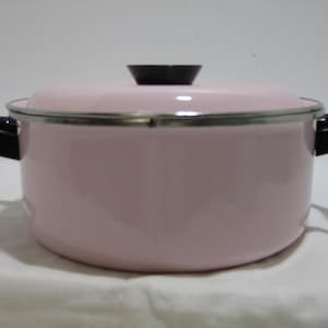 Könnte beinhalten: Ein hellrosa emaillierter Kochtopf mit Deckel. Der Topf hat schwarze Griffe und einen schwarzen Knopf auf dem Deckel. Der Rand des Topfes und des Deckels ist silberfarben. Der Topf steht auf einer weißen Oberfläche.