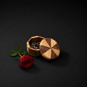 Puede incluir: Un joyero redondo de madera con un patrón geométrico de estrella en tonos de madera clara y oscura. La tapa está parcialmente abierta, revelando varios anillos de metal en su interior. Una rosa roja vibrante con hojas verdes yace al lado.