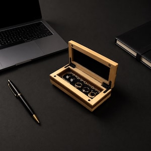 May include: A rectangular wooden jewelry box with a hinged lid, featuring a light wood exterior with a dark wood inlay. The interior is lined with black fabric and contains a gold-toned chain, two small stud earrings, and two gold rings.
