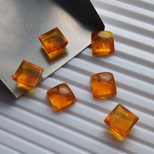 May include: Six translucent, square-cut amber gemstones with a warm orange hue are arranged on a white, ridged surface. The gemstones are slightly irregular, with visible inclusions, and are positioned near a silver metal tool.