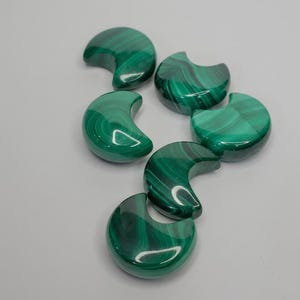 Puede incluir: Cinco piedras de malaquita pulidas talladas en forma de media luna. Cada piedra muestra un color verde intenso con patrones de bandas en espiral distintivos y una superficie lisa y reflectante dispuestas en grupo.