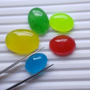 Puede incluir: Colección de cinco gemas ovaladas pulidas en colores vibrantes: verde, amarillo, rojo, azul y verde neón. Unas pinzas sostienen la gema azul. Las gemas se exhiben sobre una superficie blanca y acanalada.
