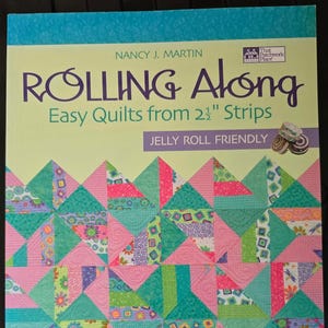 May include: A softcover instructional book titled Rolling Along Easy Quilts from 2 1/2" Strips by Nancy J. Martin. The cover features a colorful quilt pattern made of pink, teal, and patterned fabric triangles and a small jelly roll graphic.