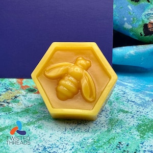 May include: A hexagonal, yellow soap bar with a raised bee design. The bee is centered within the honeycomb shape. The soap is set against a blue background and a patterned fabric.