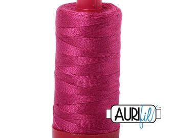 May include: A spool of vibrant magenta sewing thread featuring a smooth, cylindrical plastic core. The thread is tightly wound around the spool, which has a matching magenta top and base. The Aurifil logo is printed in the lower right corner.