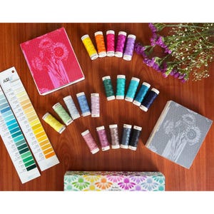 May include: A collection of sewing thread spools in various colors, two square boxes with floral patterns, a long color reference chart labeled AuriFil, and a rectangular box with a rainbow floral design, all arranged on a wooden surface.