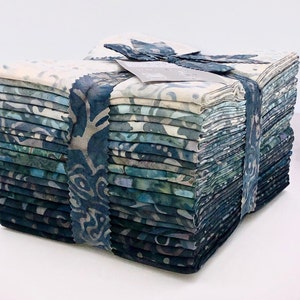 Puede incluir: Un conjunto de cuadrados de tela en tonos azules, turquesas y grises, atados con una cinta estampada. La tela presenta un diseño estilo batik, con remolinos y formas orgánicas. El conjunto está cuidadosamente dispuesto, sugiriendo un proyecto de costura.