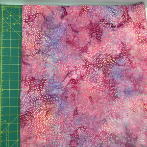 May include: A vibrant fabric with a batik-style design. The fabric features a blend of pink, purple, and blue hues, with a pattern of white, feathery shapes. The fabric is likely used for quilting or other craft projects.