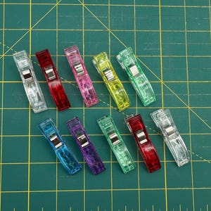 May include: A collection of colorful sewing clips in various shades, including clear, red, pink, yellow, green, blue, and purple. The clips are arranged on a green cutting mat with grid lines, showcasing their vibrant hues and functionality for fabric projects.
