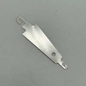 May include: A silver-colored metal knitting tool with a hook at one end and a small hole near the other. The tool has a flat, elongated shape with a notched edge.