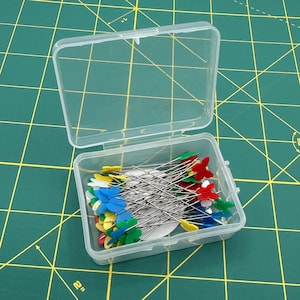 May include: A clear plastic container holding a collection of sewing pins. The pins have silver shafts and colorful flower-shaped heads in red, yellow, blue, and green. The container is open, revealing the contents on a green cutting mat.