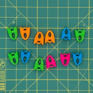 May include: A collection of colorful plastic clips in various shades of green, orange, blue, and pink. The clips are arranged on a green cutting mat with grid lines. The clips have a unique design with a central opening and curved edges.