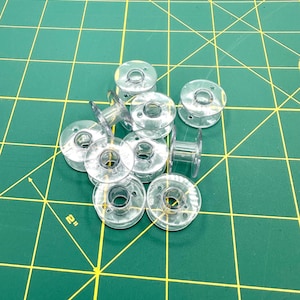 May include: A collection of clear plastic sewing machine bobbins. The bobbins are circular with a central hole and a raised edge for thread. They are scattered on a green cutting mat with yellow grid lines. The mat has a 2-inch measurement indicator.