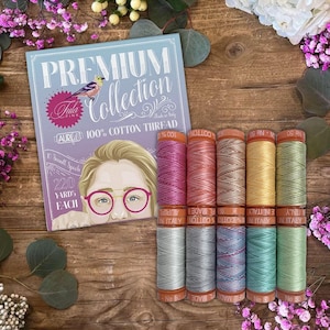 May include: A set of ten small spools of 100% cotton thread from the Aurifil Premium Collection by Tula Pink. The packaging features a blue background with a bird illustration and a portrait of a person wearing pink glasses, containing 220 yards each.