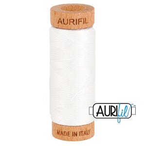 May include: A spool of white sewing thread wrapped around a cylindrical wooden core. The top of the wooden spool is branded with the text AURIFIL, and the bottom is branded with the text MADE IN ITALY. The thread has a smooth, matte finish.