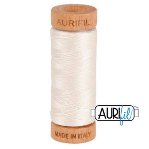 May include: A spool of off-white sewing thread wound around a cylindrical wooden core. The top of the wooden spool is branded with the text AURIFIL, and the bottom is branded with the text MADE IN ITALY. The thread has a smooth, light texture.