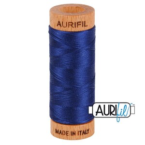 May include: A spool of deep blue sewing thread wrapped around a light-colored wooden core. The top of the wooden spool is branded with the text AURIFIL, and the bottom is stamped with the text MADE IN ITALY. A logo with the text AURIFIL is visible.