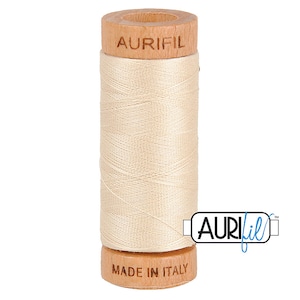 May include: A spool of Aurifil thread in a light beige color. The spool is made of wood and has the Aurifil logo printed on it. The spool is labeled "Made in Italy".