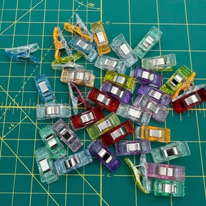 May include: A collection of colorful plastic clips for sewing and quilting. The clips are assorted colors, including red, orange, yellow, green, blue, purple, and clear.