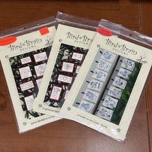 May include: Three sealed packages of Bird Brain Designs machine embroidery kits. The kits feature various designs, including Santa Redwork Ornaments and Snowmen by the Dozen. The packages have a cream-colored background with red and blue accents.