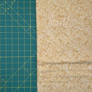 May include: A close-up of a fabric with a repeating pattern of stylized leaves in shades of tan and cream. The fabric is next to a green cutting mat with yellow grid lines and measurements in inches.
