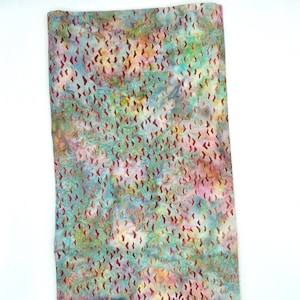 May include: A rectangular piece of fabric with a colorful batik pattern. The fabric features a blend of blue, green, pink, and orange hues, with small, reddish-brown, tear-drop shapes scattered across the surface. The fabric is likely used for sewing or crafting.
