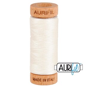 May include: A spool of white sewing thread featuring a light brown wooden top and bottom. The top wooden piece is branded with the text AURIFIL, while the bottom wooden piece is branded with the text MADE IN ITALY. The thread is wound tightly.