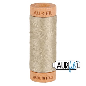 May include: A spool of Aurifil thread in a light beige color. The spool is made of wood and has the Aurifil logo printed on it. The text "Made in Italy" is printed on the bottom of the spool.