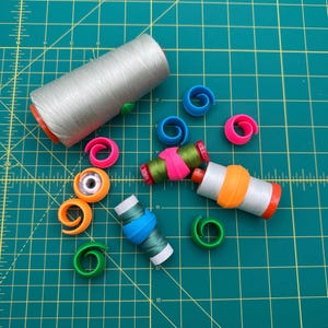 May include: A collection of sewing supplies, including spools of thread in various colors and thread spool huggers in blue, pink, orange, and green. The thread is silver, green, and white. The background is a green cutting mat.
