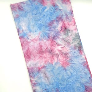 May include: A rectangular fabric with a tie-dye pattern in shades of blue, pink, and white. The fabric has a crinkled texture, with the colors blending together in a watercolor effect. The overall design is abstract and artistic.